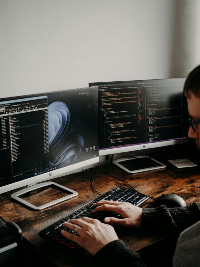 pexels-photo-16129705-16129705 A man working on website design and coding at a home office with a dual monitor setup.